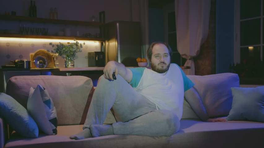 Young fat man having insomnia and zapping tv channels sitting on sofa in living room at night. Lonely obese man spending evening at home watching tv. | Shutterstock HD Video #1046488693