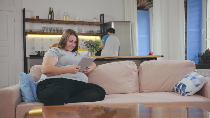 Smiling fat lady browsing internet over tablet at home. Woman holding computer at home, boyfriend cooking lunch on background in modern loft apartment