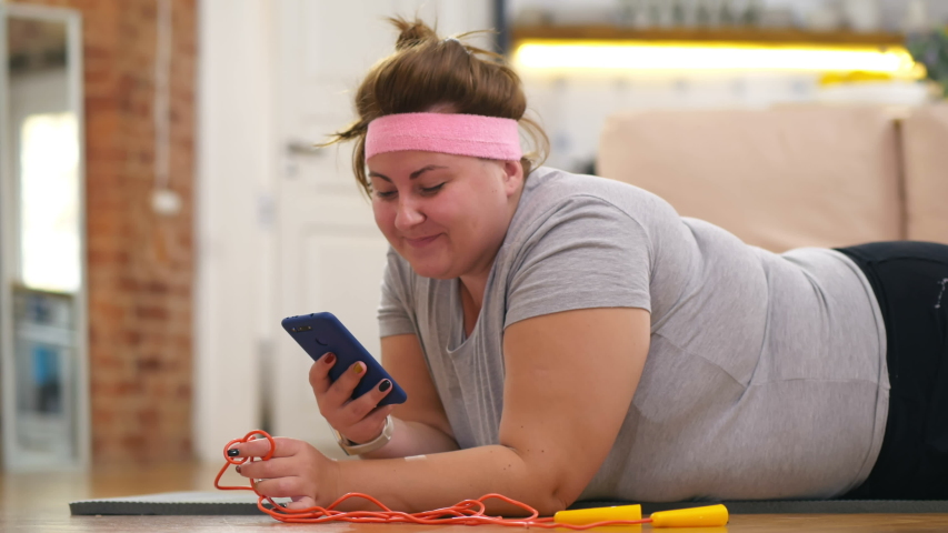 Lazy fat female lying on mat scrolling social media on gadget instead of workout. Overweight woman browsing internet oh phone instead of doing exercises. Royalty-Free Stock Footage #1046488774