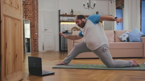 fat man doing gymnastics for losing weight at home on mat on floor watching tutorial video on laptop. Overweight guy doing yoga exercises at home.