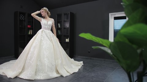 Young blonde bride in white wedding dress with crown poses for camera in room with black walls. Green leaves of houseplant are blurred in foreground