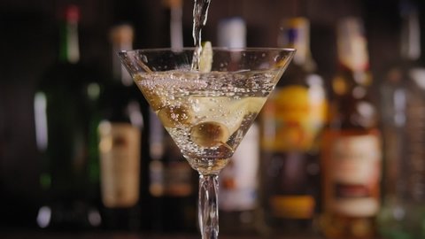 Close-up of a Martini glass with olives and lime poured Martini or gin on the bar on a blurred background of bottles of various alcohol. Slow motion. The glass turns slowly.