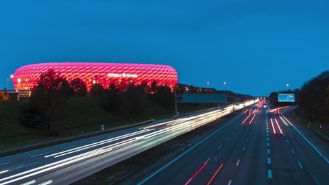 Munich / Germany - 11 17 2019: Timelapse of Allianz Arena illuminated in red neon light with highway traffic day to night. Stadium is the home arena of german soccer club FC Bayern Munich.