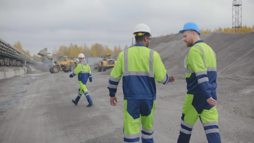 Back view of two industrial workers greeting each other and walking on construction site. Loader truck loading crushed stone in wagons of freight train. Heavy industry mining factory Royalty-Free Stock Footage #1046535292