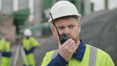 Close portrait of male builder in hardhat with walkie talkie or radio at construction site. Foreman wearing safety uniform and hardhat talking via walkie-talkie while standing at construction site