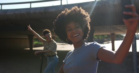 Front view of two mixed race women enjoying free time in a urban park on a sunny day together, riding electric scooters, taking photos, slow motion