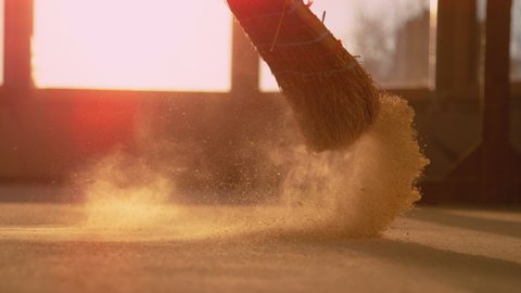 SLOW MOTION, LENS FLARE, CLOSE UP, LOW ANGLE: Dust gets swept up into air as an unrecognizable person cleans a construction site. Contractor sweeps the ground of a construction site with a straw broom