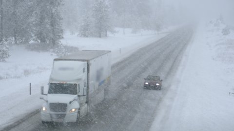 Semi-truck and car drive along snowy country road in horrible weather conditions. Two vehicles carefully navigate the interstate road during a blizzard. People driving across state in poor visibility.