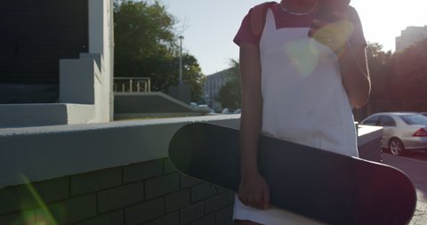 Front view of mixed race woman enjoying free time on a street on a sunny day, standing, using smartphone and holding a skateboard, slow motion