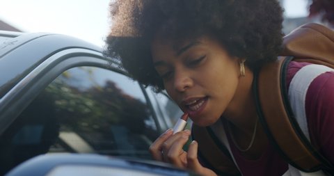Front view close up of mixed race woman enjoying free time on the street on a sunny day, putting lipstick on, looking in a car mirror, slow motion