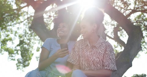 Low angle front view of two mixed race women enjoying free time in a urban park on a sunny day together, using smartphone, sharing earphones and listening to music, slow motion