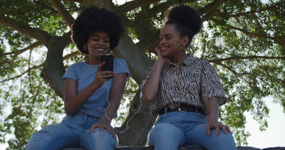 Low angle front view of two mixed race women enjoying free time in a urban park on a sunny day together, using smartphone, sharing earphones and listening to music, slow motion