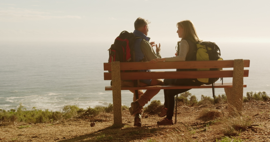 Side view of a senior Caucasian couple enjoying time in nature together, sitting on a bench on a cliff during a hike, talking in slow motion