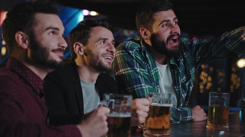 In a pub good friends drinks beer from a beer glass and watching concentrated favorite match on tv they screaming and are very excited while watching a TV game
