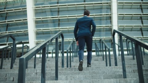 Back view stylish business man going upstairs in suit at street. Successful businessman walking at stairs alone outdoors. Young man walking in luxury suit near stadium.