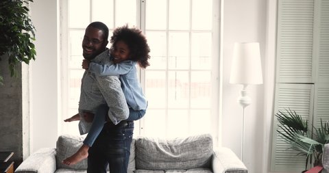 Happy african dad carrying preschool kid daughter give piggyback ride at home, loving young adult afro american father play funny active game with small child girl having fun laughing in living room