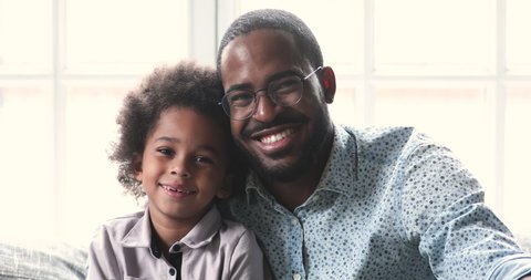 Cheerful smiling african american young adult father and little cute kid son looking at camera laughing at home, mixed race family dad child boy happy faces sit on sofa bonding, closeup portrait