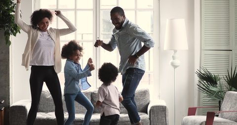 Happy carefree african american ethnic couple dancing with cute active preschool small children in living room interior together, family mortgage and parents with kids weekend fun at home concept
