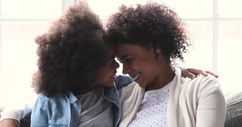 Loving happy african american family young adult mum and cute preschooler mixed race child kid daughter laughing cuddle having fun sit on sofa look at camera enjoy sweet moment play at home together