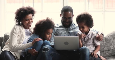 Happy relaxed african american parents with two little kids enjoying watching videos or doing family online shopping looking at laptop screen using notebook technology sit on sofa in living room