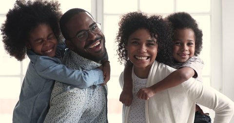 Happy african american family young adult parents father mother piggyback small cute mixed race children son and daughter laugh playing together in living room look at camera stand at home, portrait