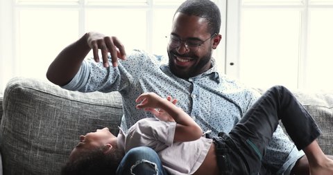 Funny happy young adult african american daddy tickling cute small son relaxing on sofa, loving ethnic father having fun laughing playing with little adorable cuddling together sit on couch at home