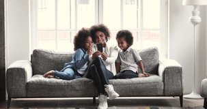 Happy young african mum using social media app on smartphone recording video blog watching stories listening music having fun with two cute small kids looking at mobile phone relaxing on sofa at home
