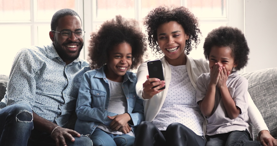Happy african family parents and small children laugh using smart phone gadget relax together on sofa, mixed race mom dad preschool kids hold look at smartphone screen use funny ar mobile app at home Royalty-Free Stock Footage #1046570521