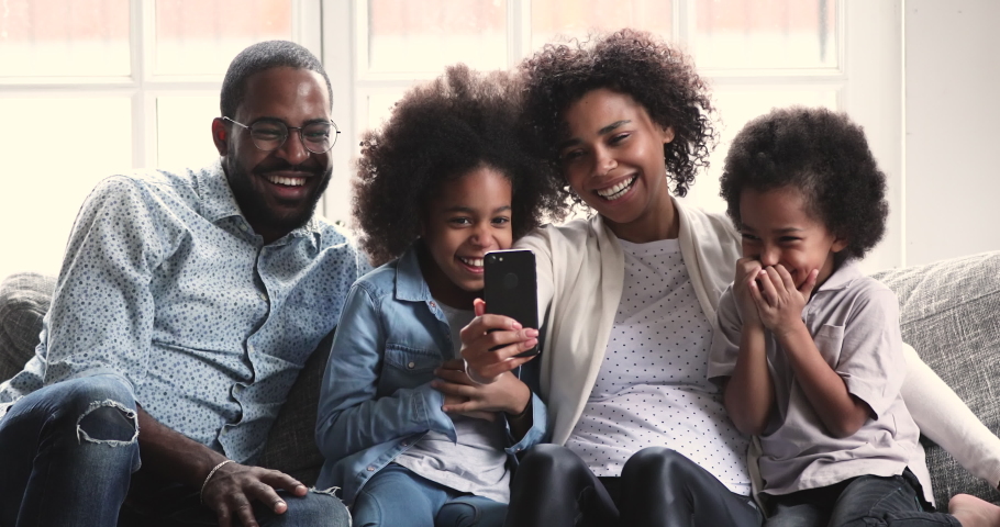 Happy african family parents and small children laugh using smart phone gadget relax together on sofa, mixed race mom dad preschool kids hold look at smartphone screen use funny ar mobile app at home
