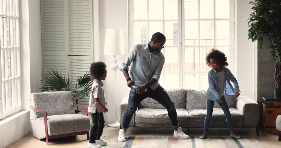 Happy active african american dad teaching dancing having fun with two cute small kids son and daughter imitate father moves playing together enjoying funny weekend activity in modern living room | Shutterstock HD Video #1046572282