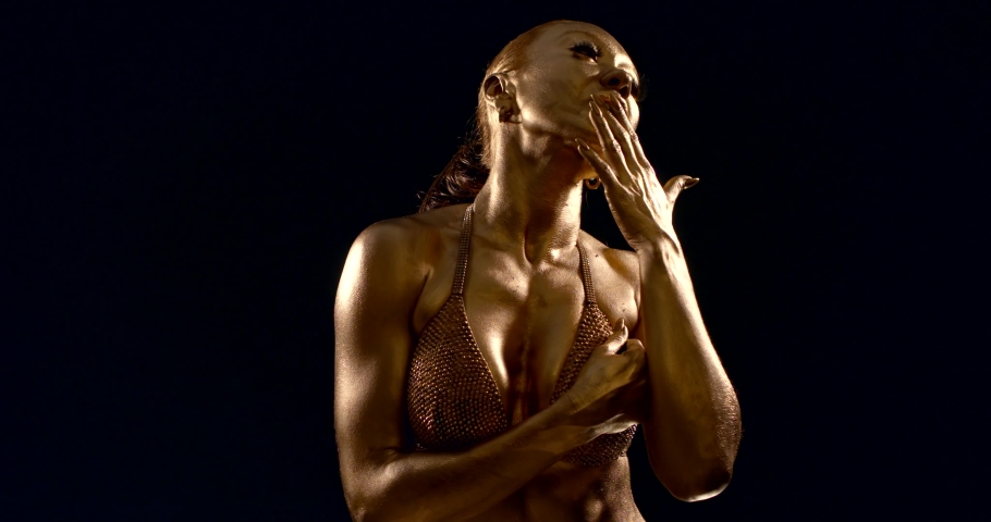 portrait of a woman with Golden skin and raised muscles in shining underwear on a black background. she runs her hand over her lips and face, looks at the camera, turns sideways, touches her hair