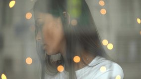 An Asian girl is sitting in a cafe alone and waiting for someone, she is sad and is thinking about something. Video from the street through the glass in a cafe.