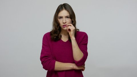 Confused thoughtful brunette woman in pullover biting lips from anxiety while pondering with perplexed expression, looking puzzled worried, having doubts. studio shot isolated on gray background