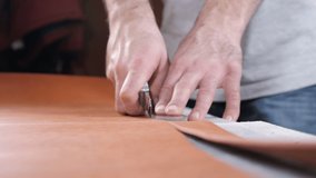 Hands of skilled leather craftsman using plastic pattern and leather knife to cut out part from piece of red leather, close-up shot. Industrial knife man cuts the work piece from the skin. 4 k footage