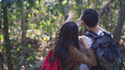 HD Slow Motion rear view of happy young Asian couple man and beautiful woman holding hands together and hiking with backpack in forest mountain with using smartphone taking a photo of nature.