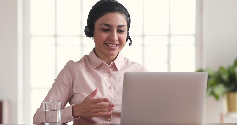 Friendly young business woman indian call center agent professional operator wear wireless headset conference calling on laptop talking to customer service support in video chat sit at desk in office