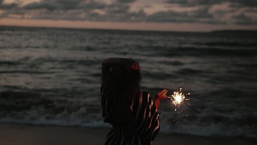 Close up portrait of young beautiful woman at sunset on the beach. Pretty girl holding sparkling fireworks in slow motion. Holiday with fireworks. City people travel. Business woman at vacation.