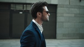 Side view serious businessman walking in suit near modern building. Confident business man wearing sunglasses at street. Portrait attractive man checking time on wrist watch outdoors.