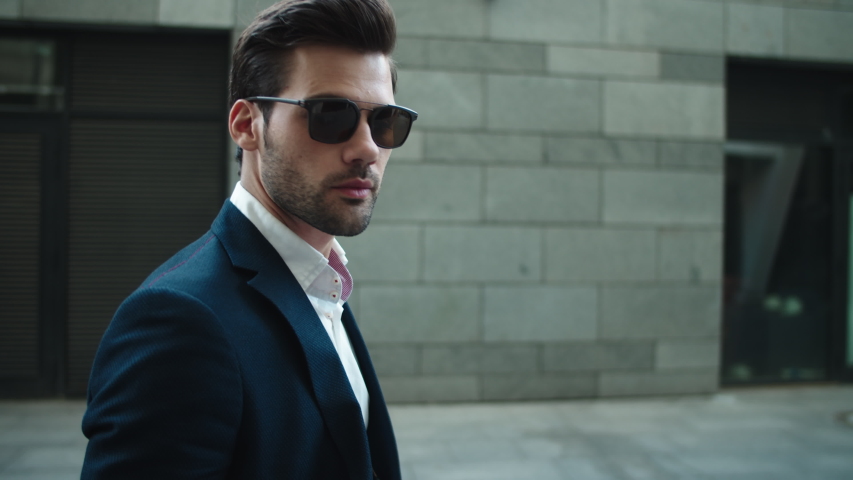 Side view serious businessman walking in suit near modern building. Confident business man wearing sunglasses at street. Portrait attractive man checking time on wrist watch outdoors. | Shutterstock HD Video #1046591341