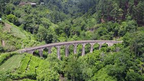 Stunning slow zoom-in 4k aerial drone stock footage of a bright red train crossing the Nine Arches Bridge. The old stone bridge is set in a green valley located in between Ella and Demodara, Sri Lanka