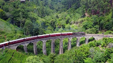 Stunning slow zoom-in 4k aerial drone stock footage of a bright red train crossing the Nine Arches Bridge. The old stone bridge is set in a green valley located in between Ella and Demodara, Sri Lanka