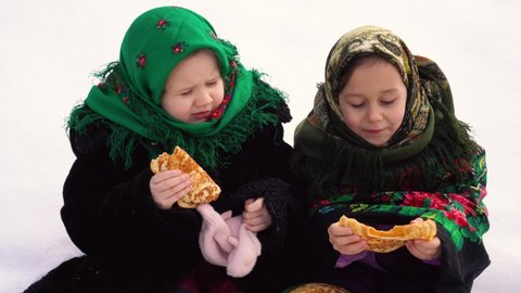 Two charming girls in a Russian folk costume sit on the snow and eat pancakes. The concept of the celebration of the Russian holiday shrovetide