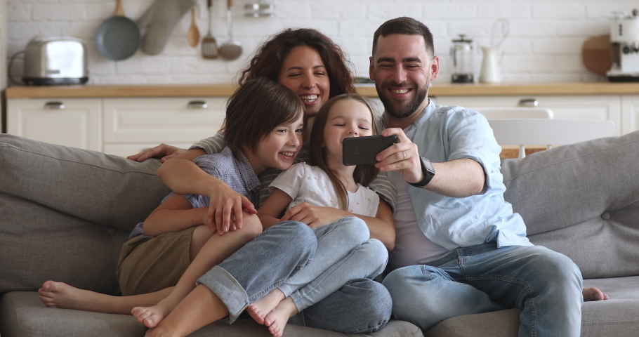 Happy bonding family of four sitting on couch, smiling father holding mobile phone, taking selfie. Excited couple posing for photo with children siblings or holding online call with grandparents. Royalty-Free Stock Footage #1046601994