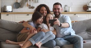 Happy bonding family of four sitting on couch, smiling father holding mobile phone, taking selfie. Excited couple posing for photo with children siblings or holding online call with grandparents.