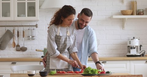 Young handsome man cuddling from back smiling attractive wife in apron preparing healthy vegetarian dish for romantic dinner. Joyful millennial woman feeding loving husband while cooking in kitchen.
