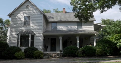 Establishing shot of a two story grey house with wood siding and a person sitting in a rocking chair on the front porch in the summer