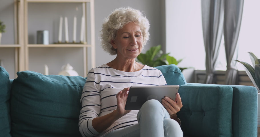 Pleasant smiling elderly mature woman resting on sofa, using digital tablet alone at home. Happy older pensioner web surfing information, chatting on social networks, studying or shopping online. Royalty-Free Stock Footage #1046611594