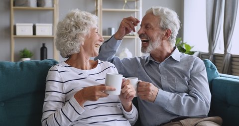 Affectionate mature family couple relaxing on cozy sofa, enjoying pleasant conversation, holding cups of hot tea. Loving happy middle aged man joking talking discussing with elderly smiling wife.