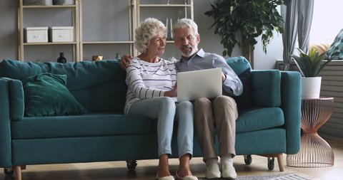 Full length happy older family couple relaxing on comfortable couch at home, using computer. Smiling middle aged married retired spouses web surfing, discussing news, shopping online together.