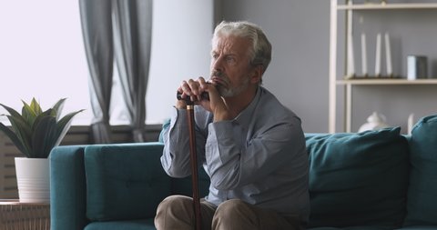 Thoughtful tired middle aged man sitting on couch relying on walking stick, feeling stressed. Depressed mature grandfather thinking of geriatric health problems disease, suffering from loneliness.
